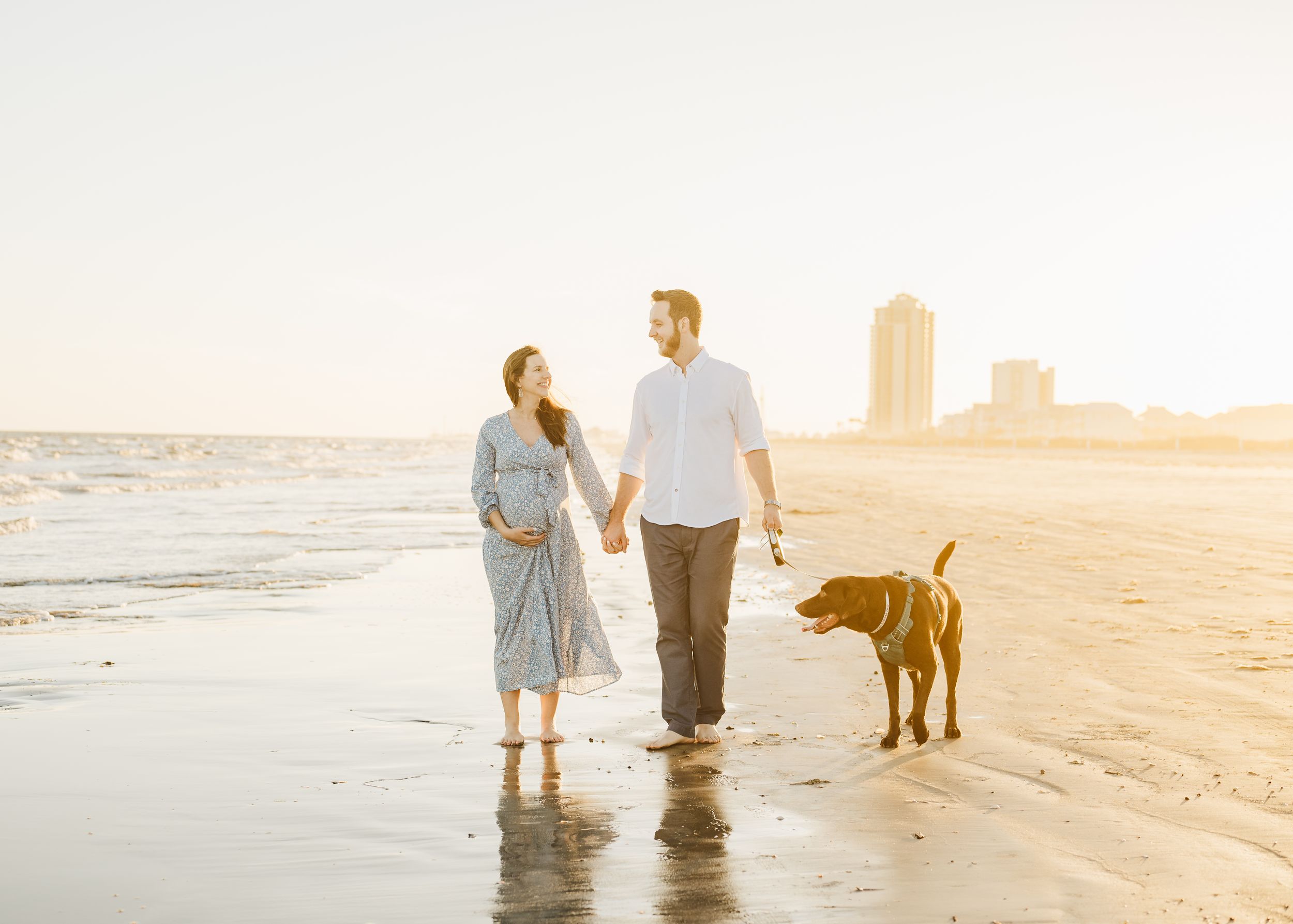 Couple and dog walking on beach at sunset with soft golden light.