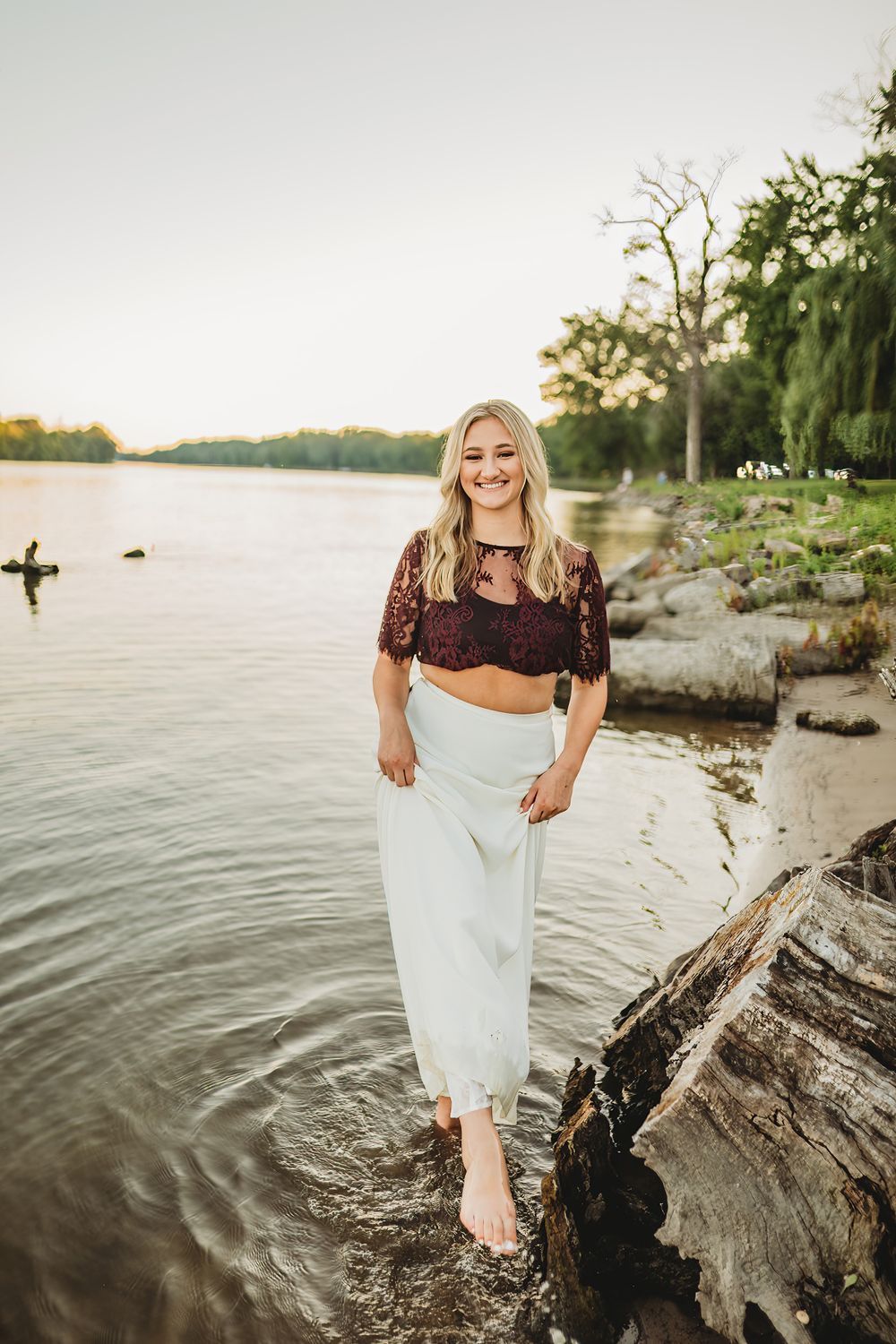 Person in white dress poses on driftwood by a serene waterfront at sunset.
