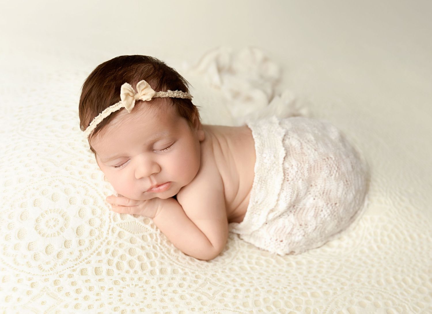 A sleeping newborn baby girl draped in white lace with a gold bow headband poses on a cream textured blanket.
