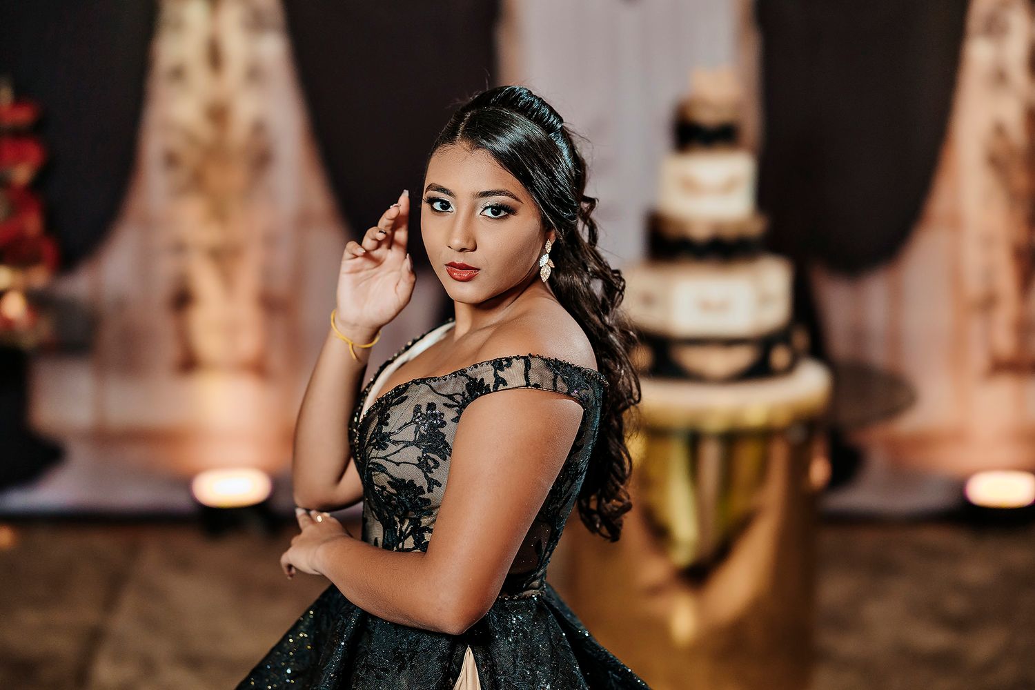 Elegant figure in a dark formal gown poses gracefully at an event with a tiered cake and decorative backdrop.