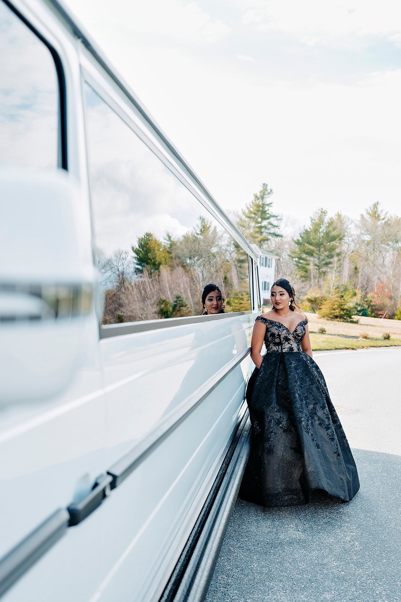 Elegant black formal gown reflected in the side of a white limousine with trees in background.