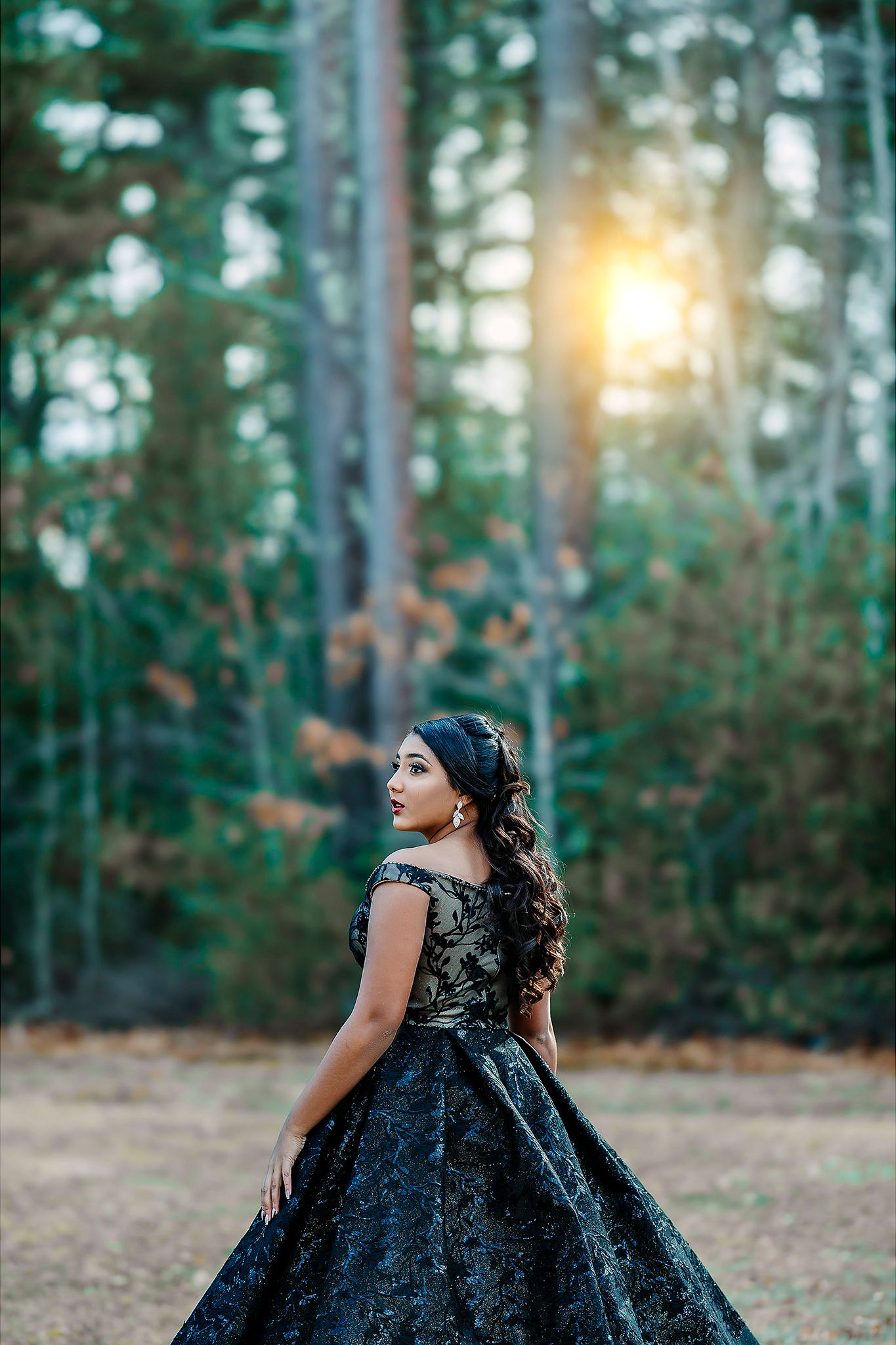 Woman in elegant black lace gown poses gracefully in a serene forest setting during golden hour.