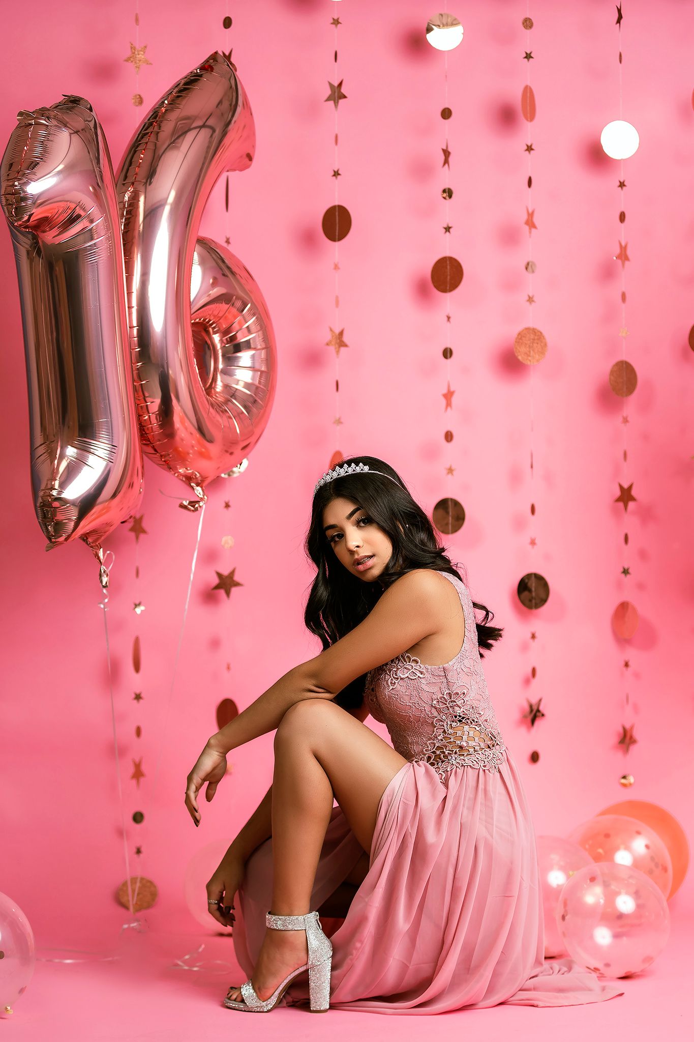 A figure in a sparkly pink dress poses with silver balloon decorations against a pink backdrop with hanging decorations.