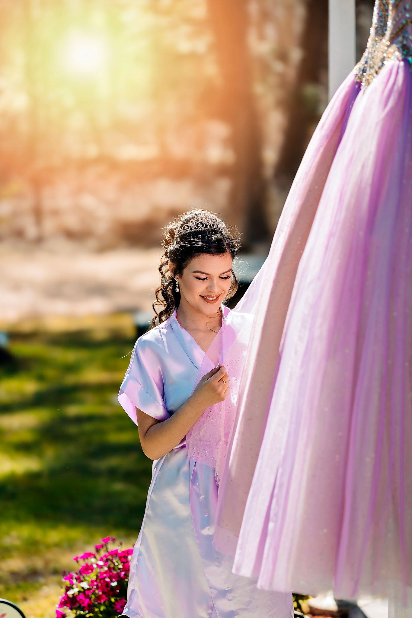 A woman in a white satin robe admires a pink tulle dress hanging outdoors in golden sunlight.