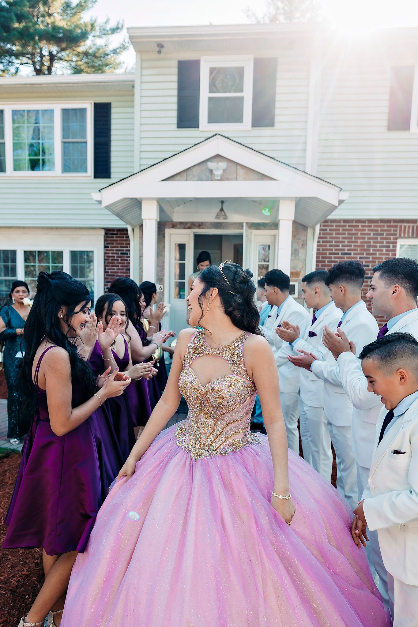 A quinceañera celebration with guests in purple dresses and white suits dancing outside a white house.