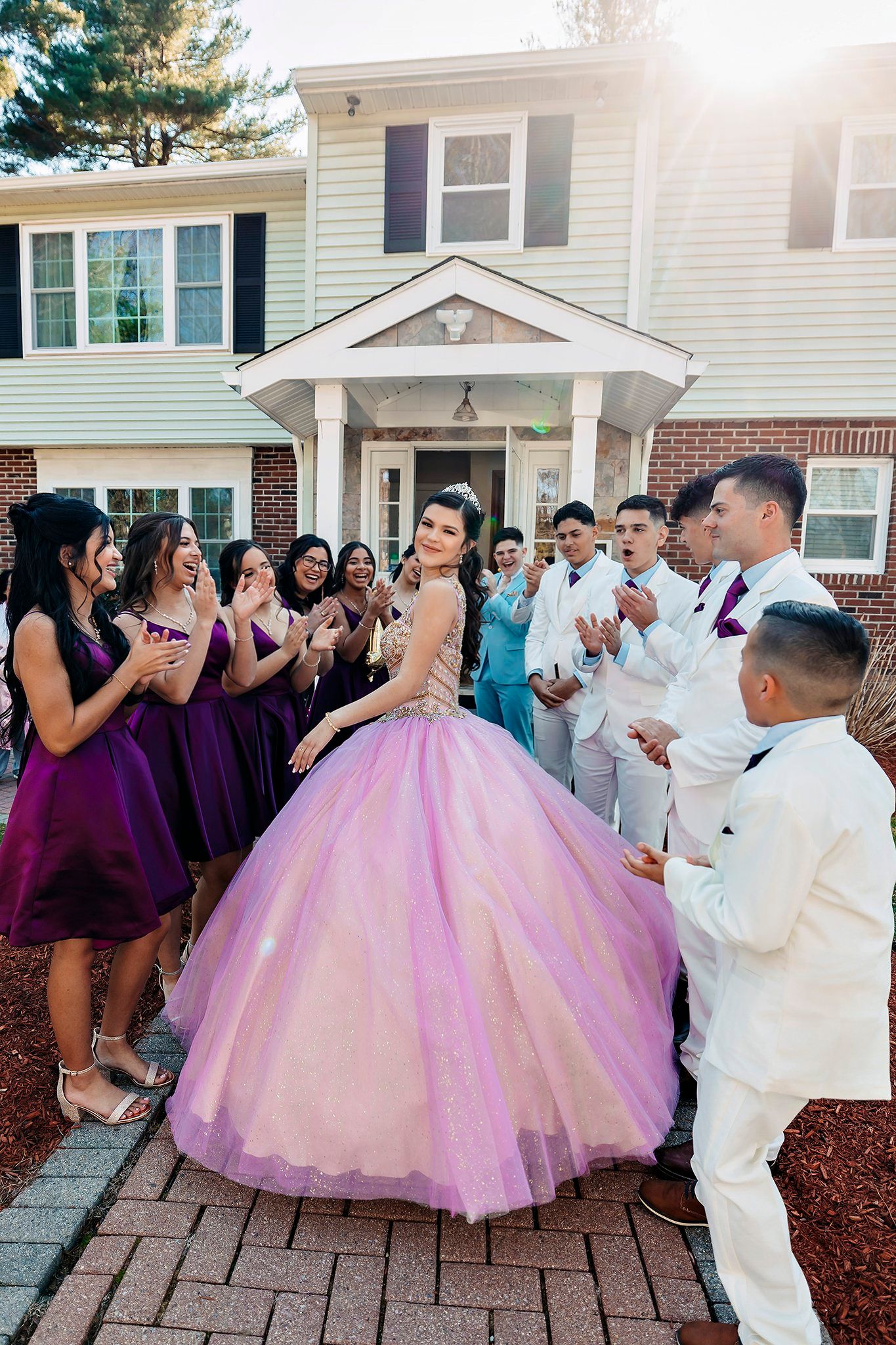 A quinceañera celebration with guests in purple dresses and white suits gathered on a brick pathway outside a house.