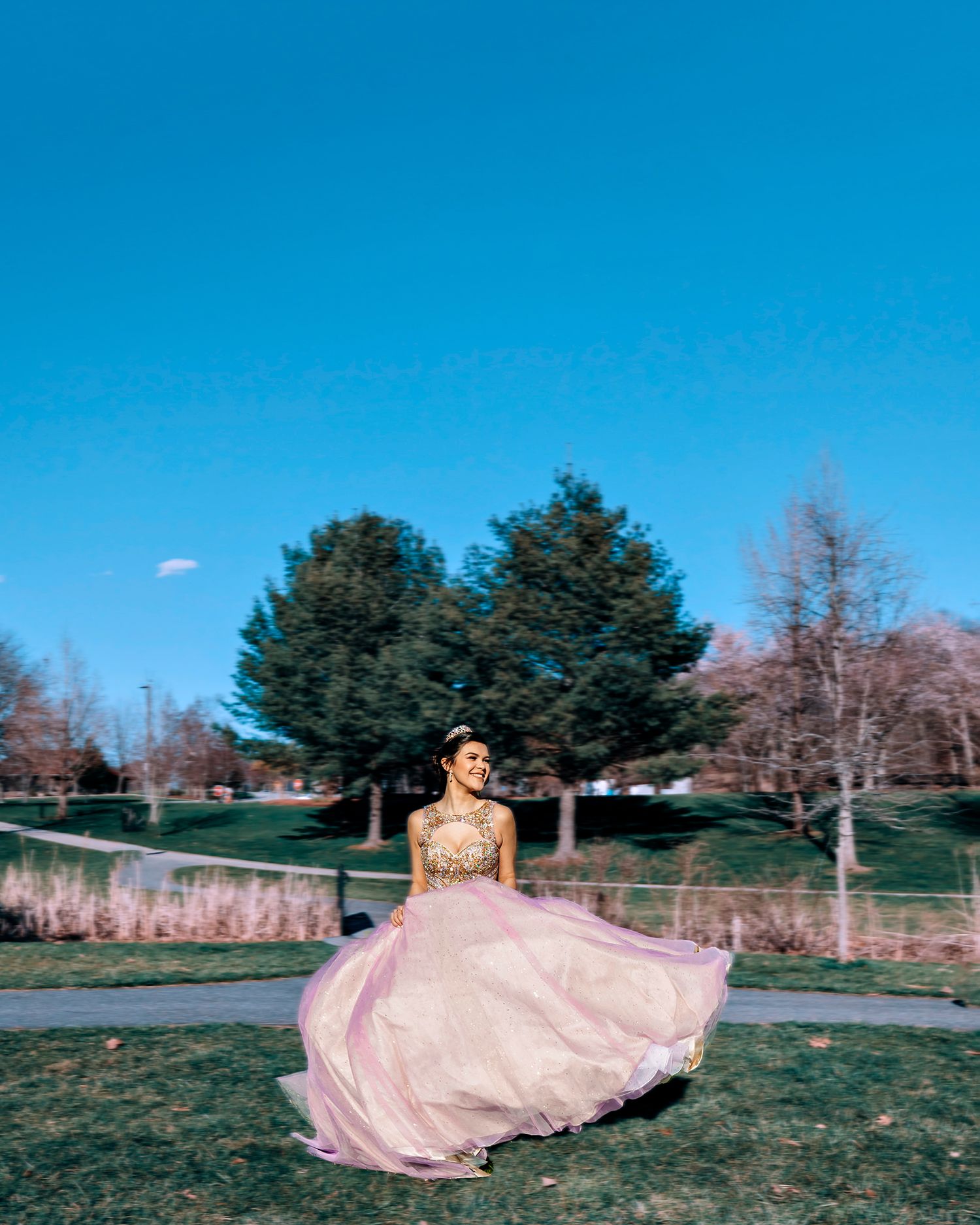 Someone in a sparkly pink gown twirls outdoors on a sunny day with evergreen trees and blue sky in the background.