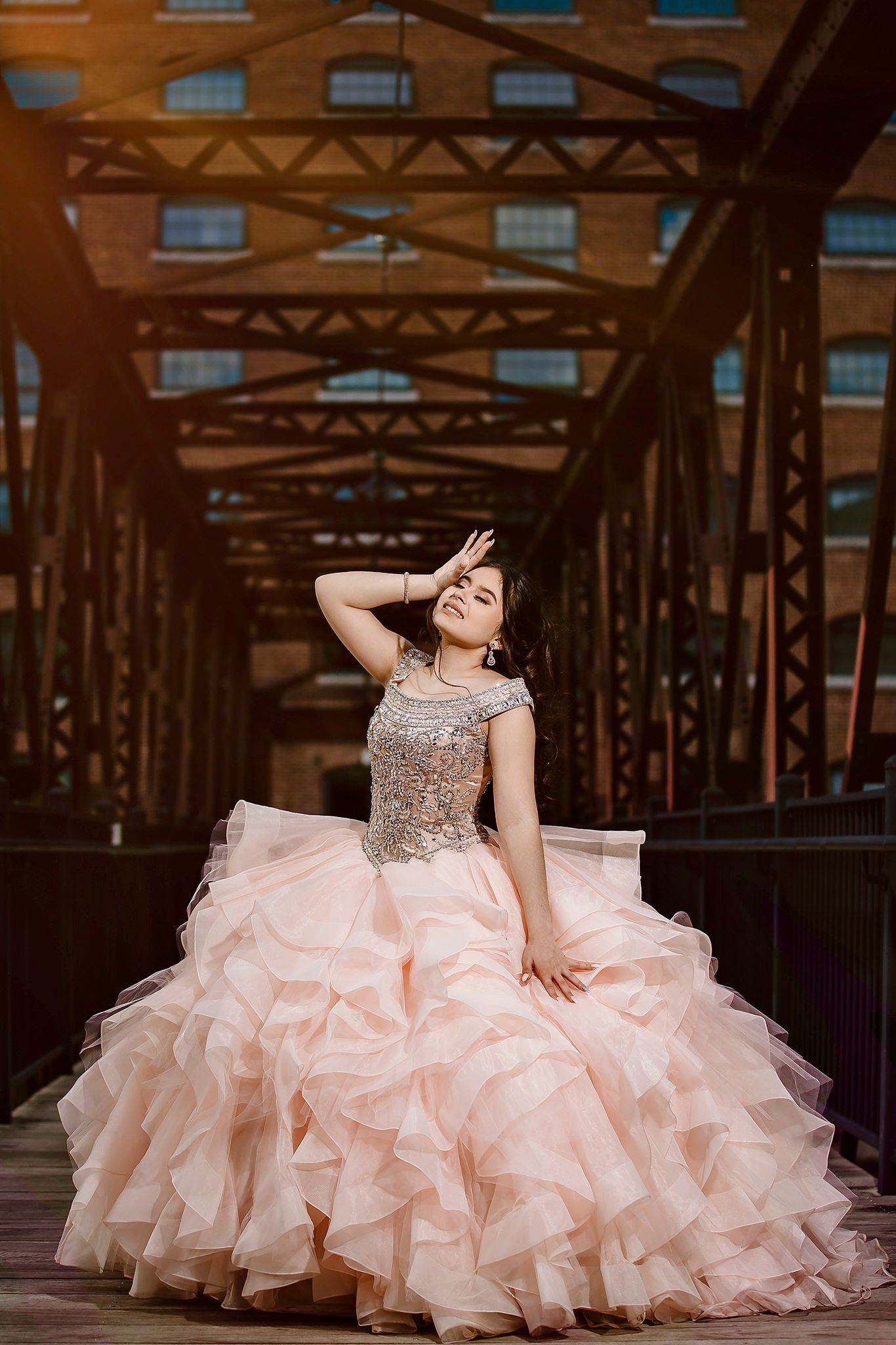 A person in a blush pink layered ballgown with crystal bodice poses gracefully on a rustic bridge.