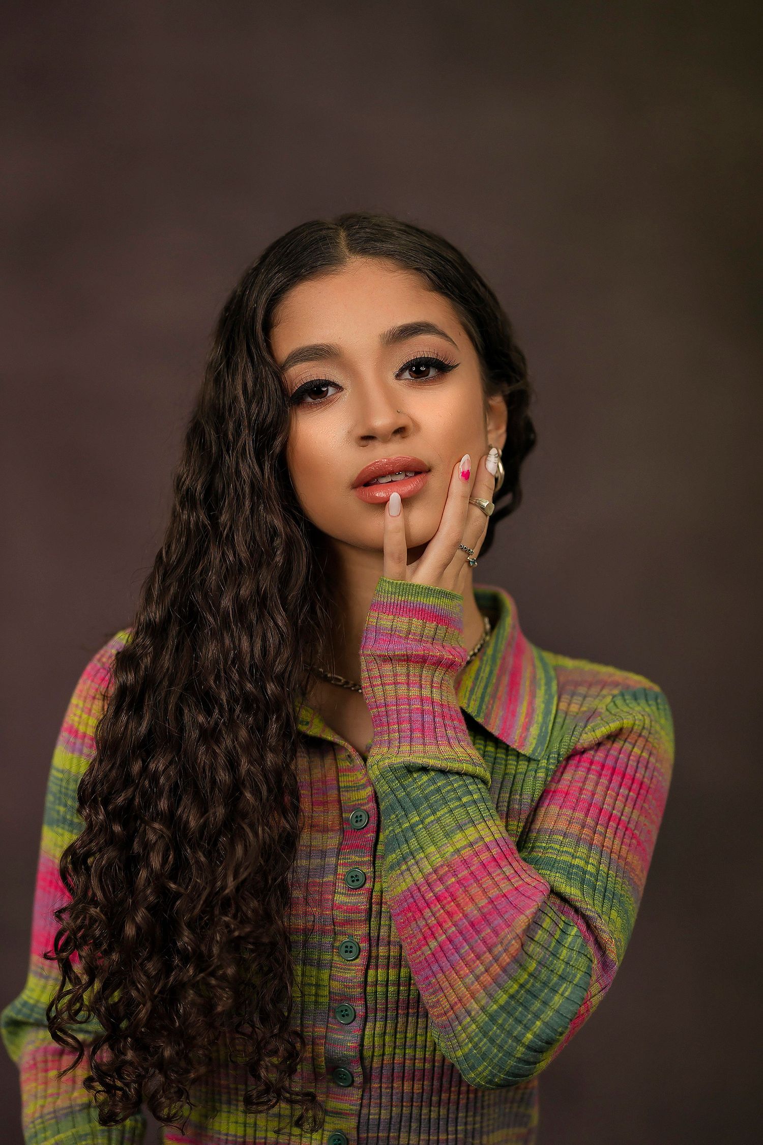 Studio portrait with long curly hair wearing a colorful striped knit sweater in pink and green against dark background.