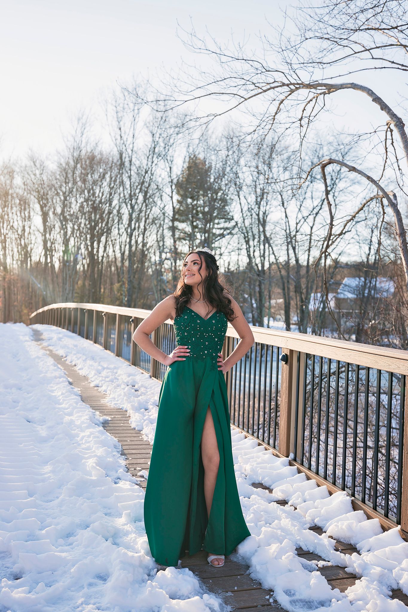 A woman in an emerald green gown poses on a snowy footbridge surrounded by bare winter trees.
