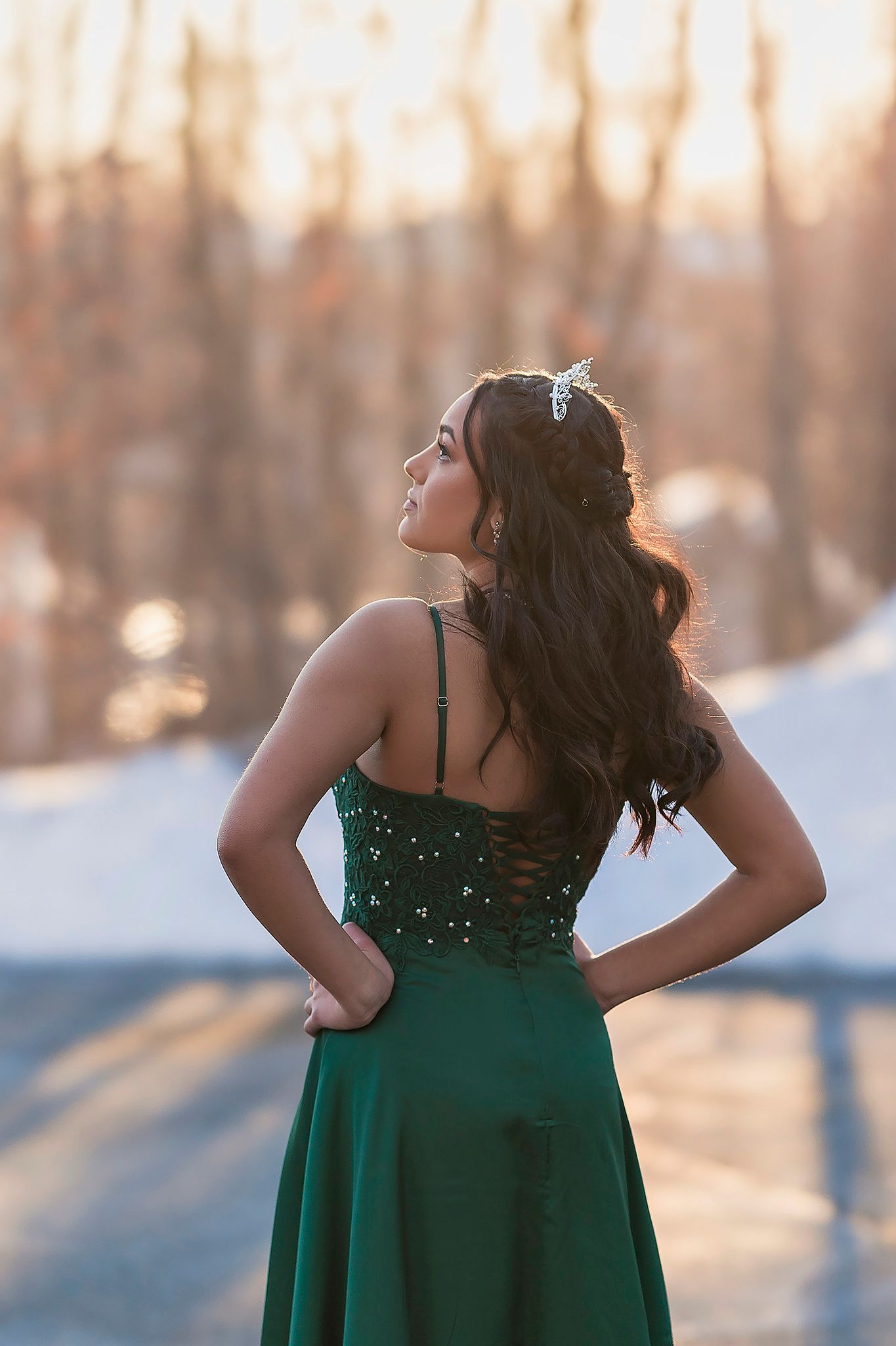 Woman in emerald green formal dress stands with back turned against snowy winter landscape at golden hour.