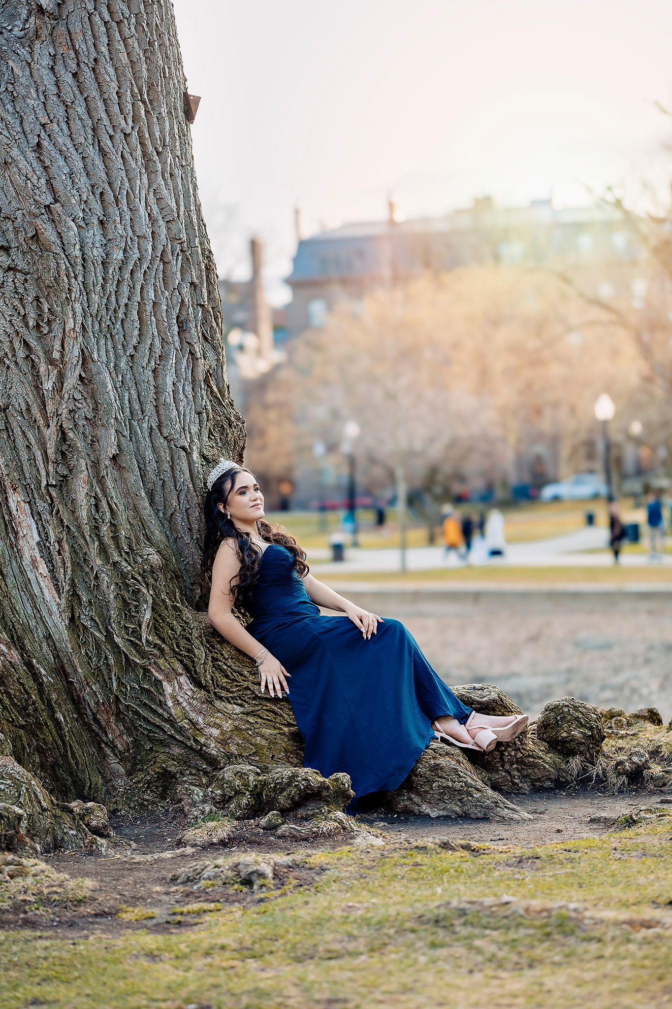 A person in a long blue dress rests against a large tree trunk in a park setting during early spring.