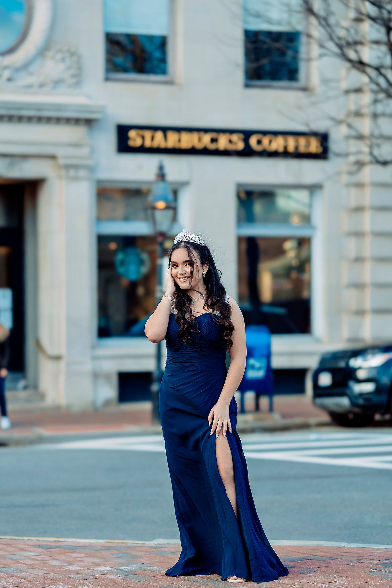 A woman in a navy blue evening gown and tiara poses on a brick sidewalk in front of a Starbucks Coffee store.