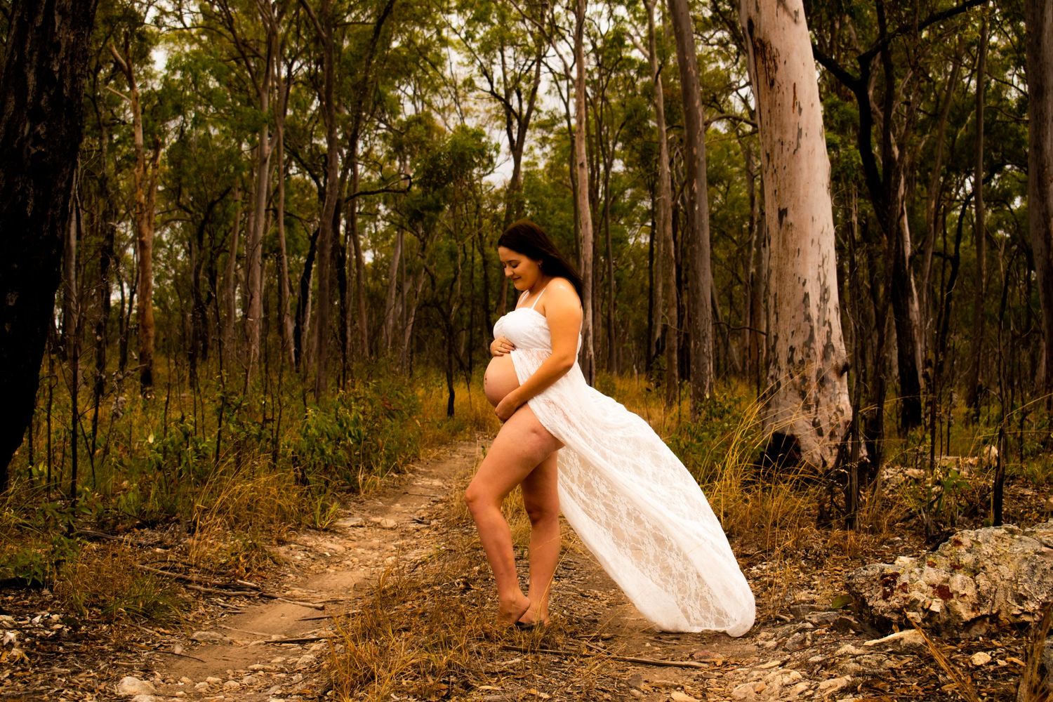 Maternity photo of woman in white dress on forest path.