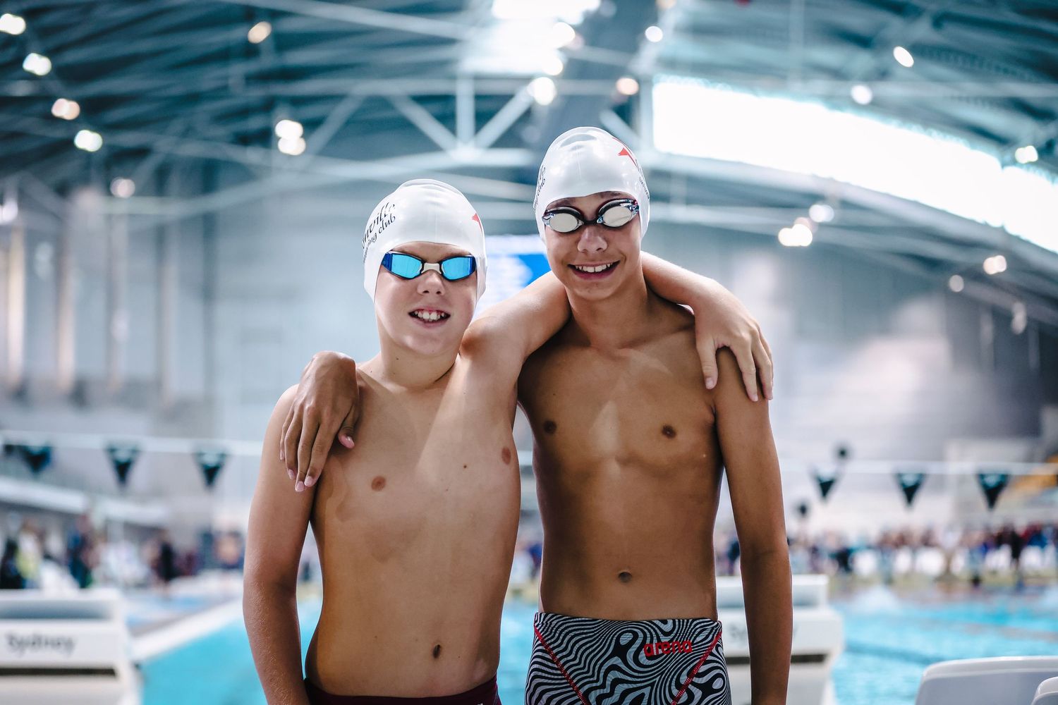 Two swimmers in goggles and swim caps posing together at indoor pool.