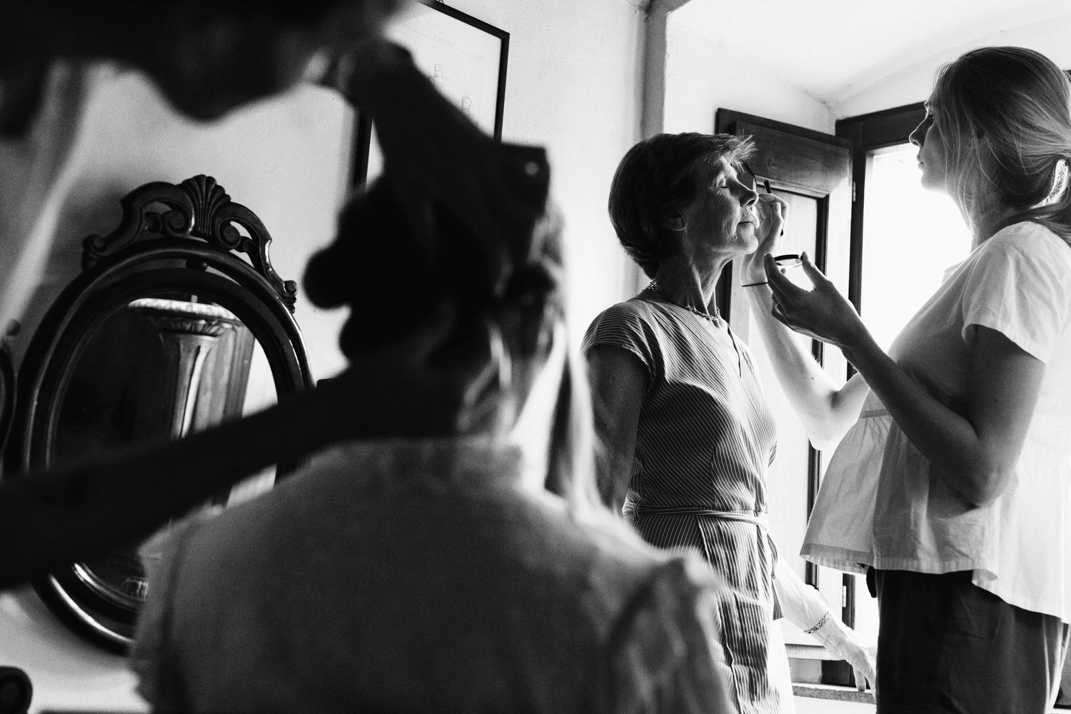 Black and white image of wedding preparation with stylists working in natural window light.