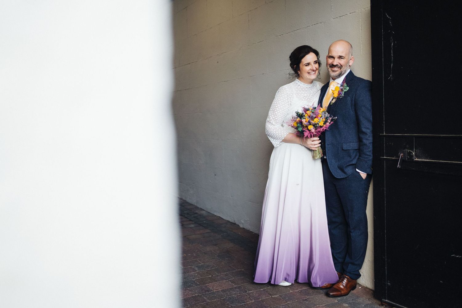 Couple poses in doorway wearing wedding attire with bride holding vibrant bouquet.