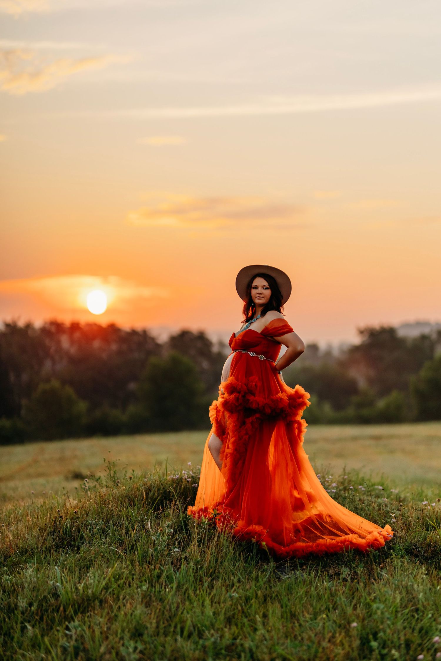 Pregnant woman in flowing red gown and hat stands in a field at sunset.