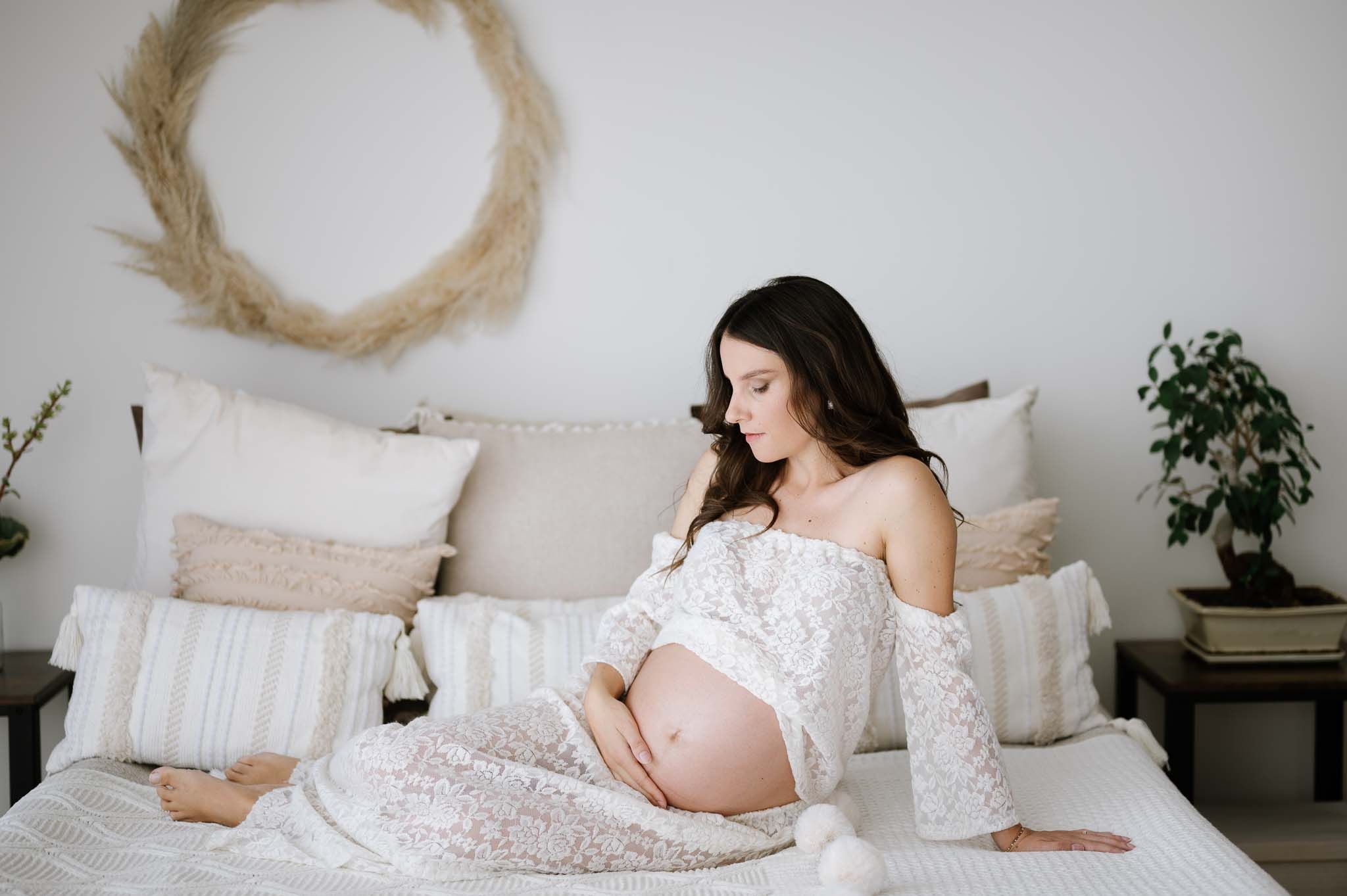 Expectant mother in white lace dress sits serenely on bed with neutral decor.