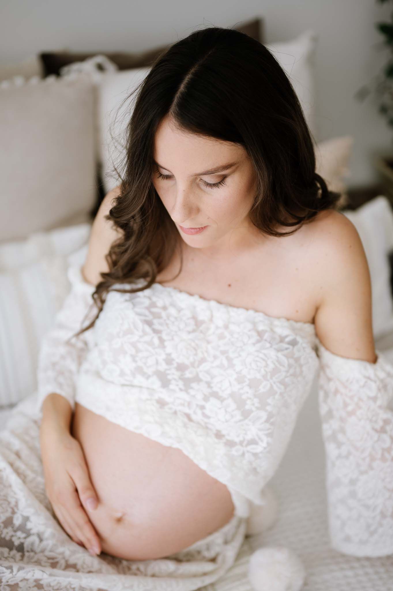 Intimate maternity portrait in white lace dress against soft neutral background.