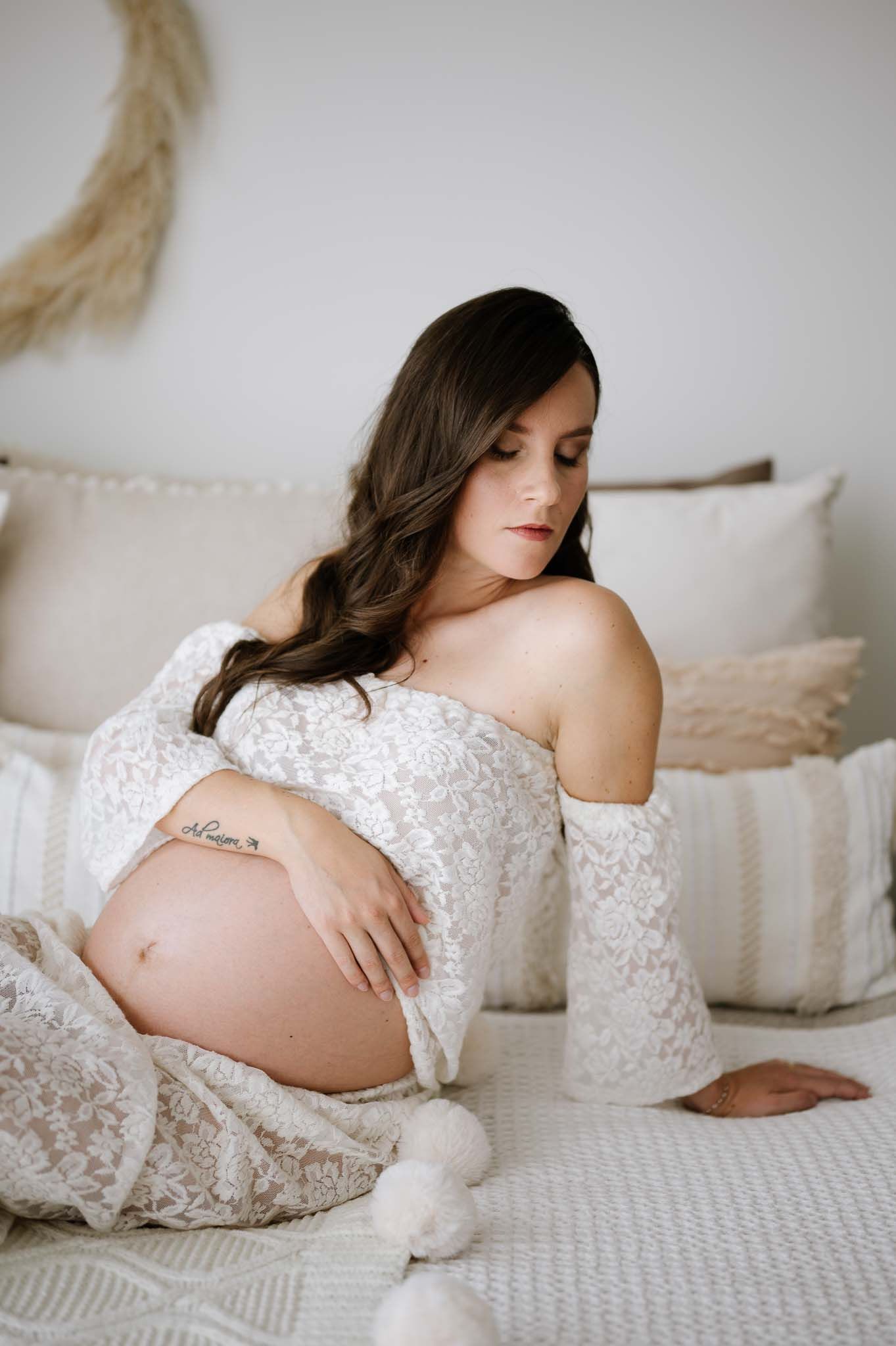 Intimate maternity photo featuring white lace dress and soft bedroom setting with decorative pillows.