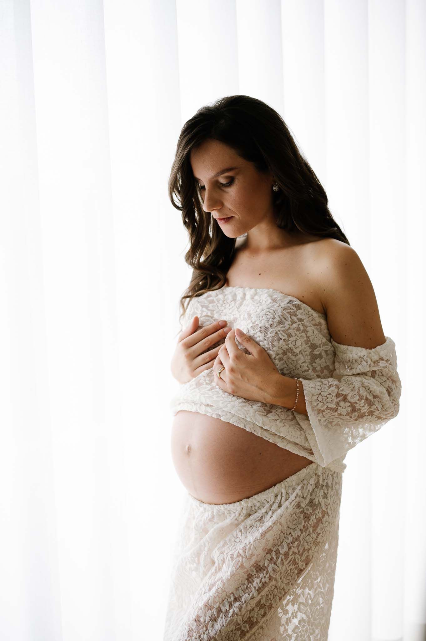 Dreamy side profile maternity photo with white lace dress against bright window light.
