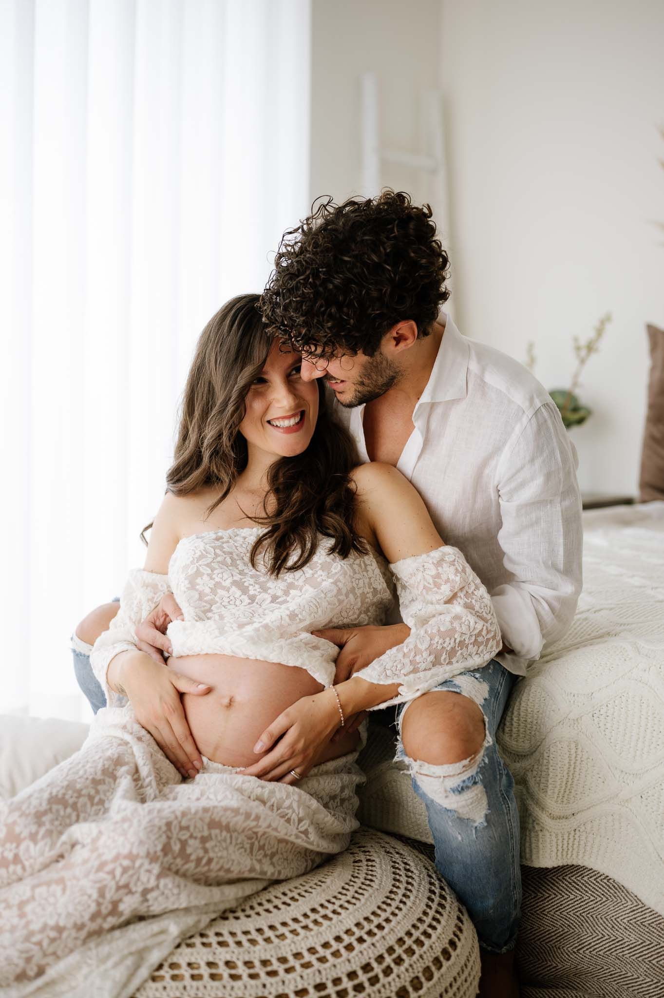 Intimate couple's maternity photo on couch with decorative boho elements and natural lighting.