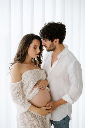 Intimate maternity portrait against white curtains with soft natural lighting and tender moment.