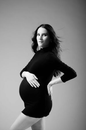 Black and white maternity photo of woman in short turtleneck dress with windswept hair against plain backdrop.