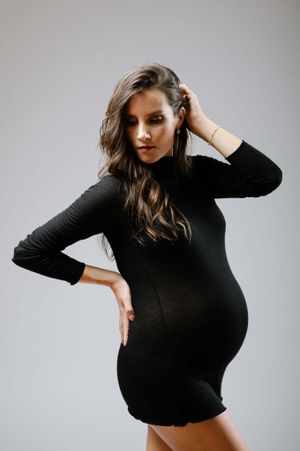 Expectant mother poses in fitted black dress showing silhouette against grey background.