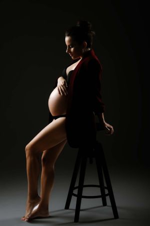 Silhouetted maternity photo of a pregnant woman in a red jacket sitting on a stool against dark background.