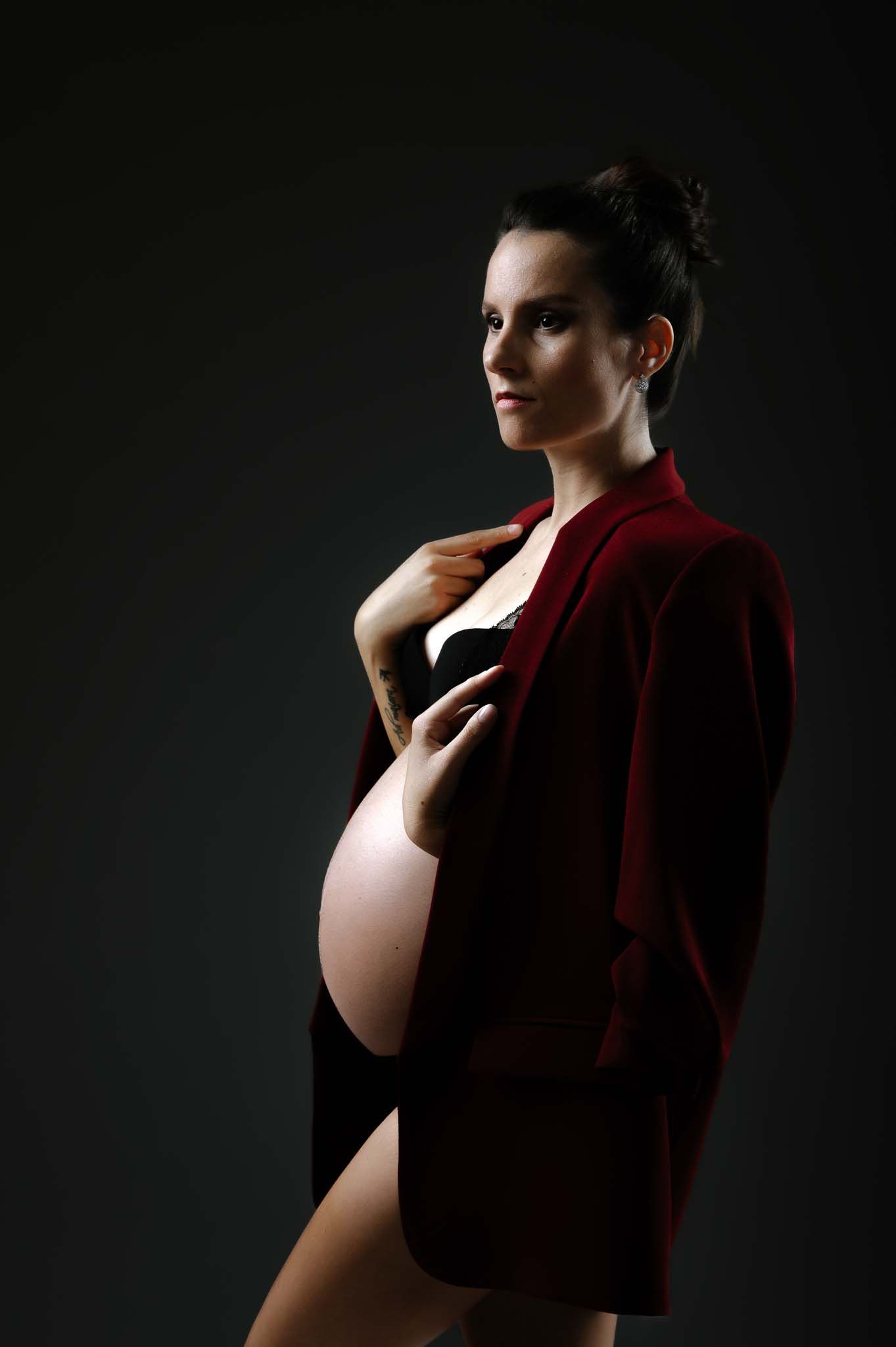 Artistic maternity photo featuring pregnant figure in red jacket against dark backdrop.