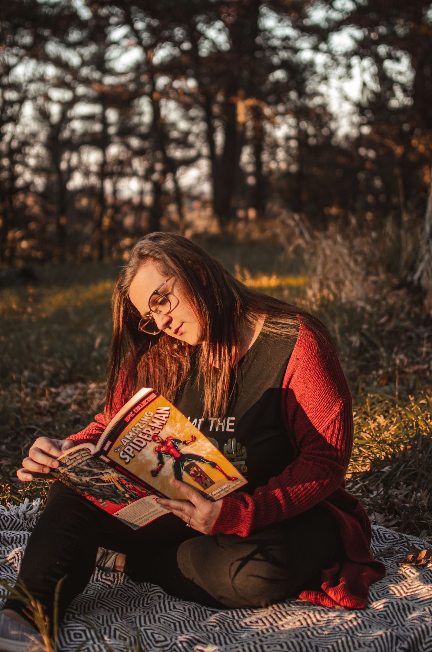 Person reading a book while sitting on a patterned blanket in an autumn forest setting.