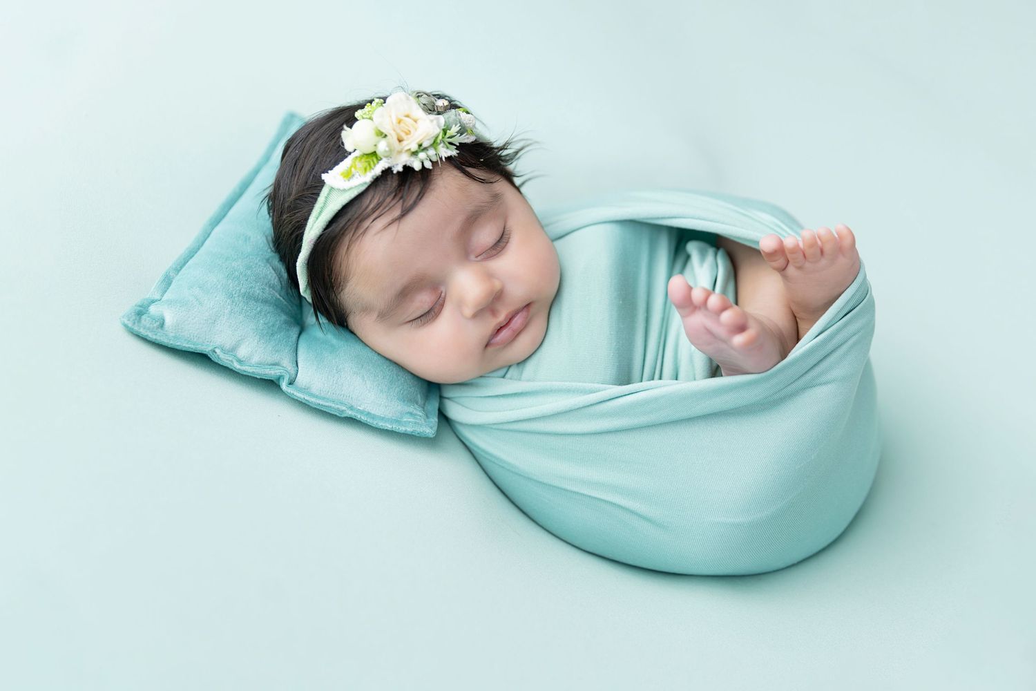 Sleeping newborn wrapped in turquoise fabric with floral headband.