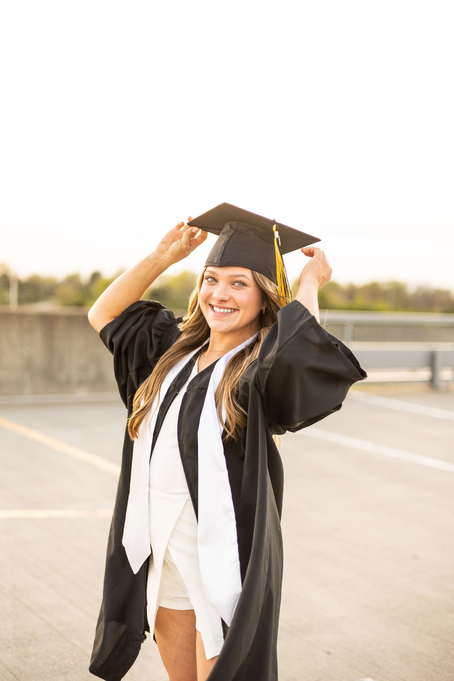 Smiling graduate in black cap and gown adjusting tassel outdoors on rooftop at golden hour.