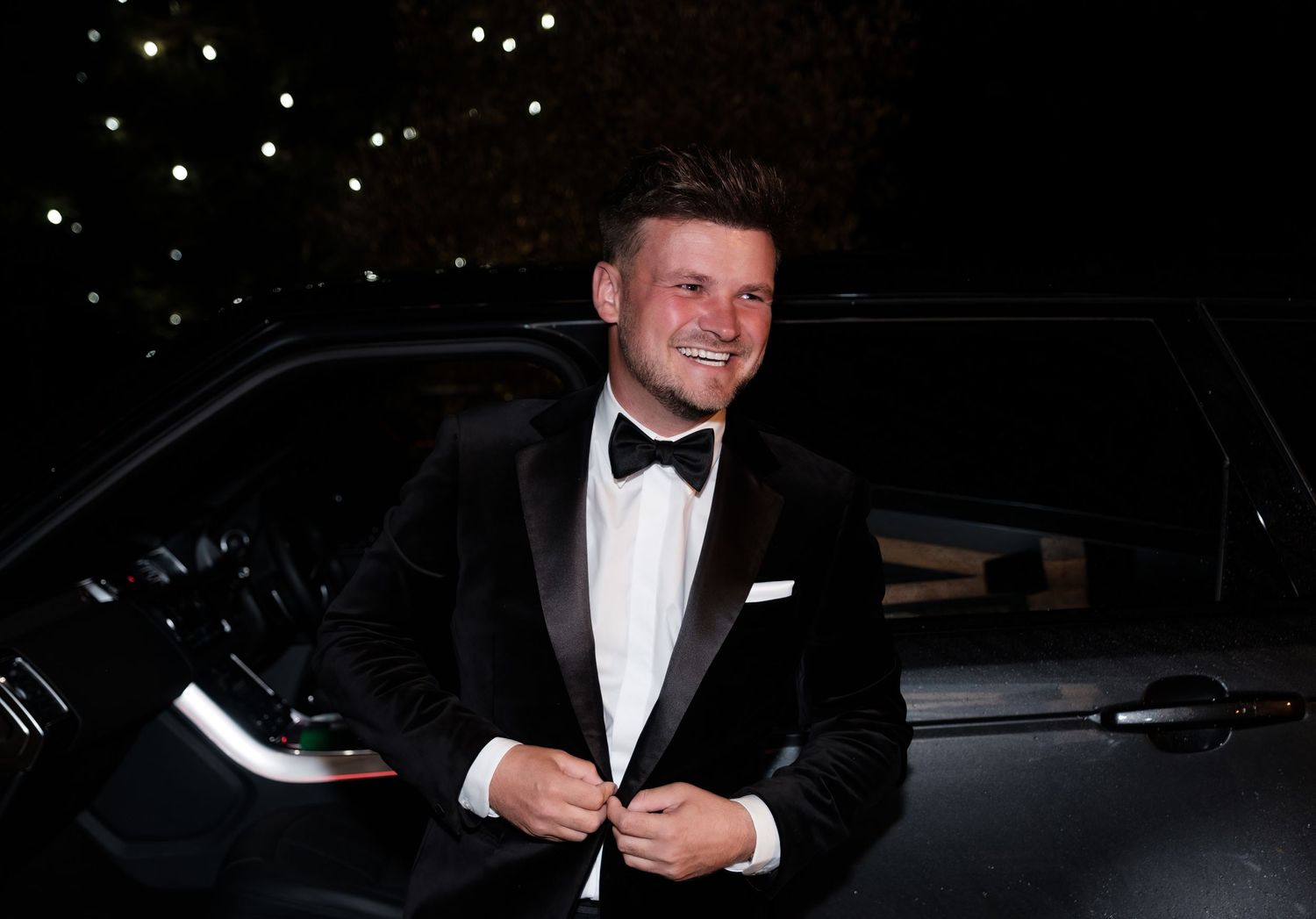 An elegant groom in a black tuxedo and bowtie standing next to a car at night.