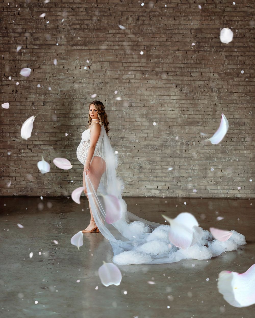 A dreamy maternity photo with floating white feathers against a brick wall backdrop.