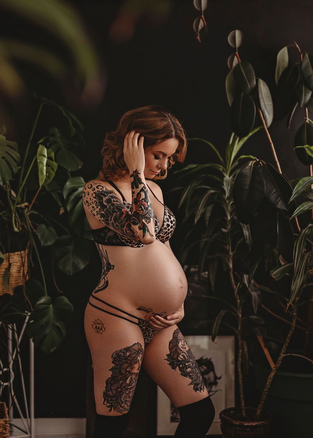 Artistic maternity photo with tattooed woman in lingerie among houseplants.