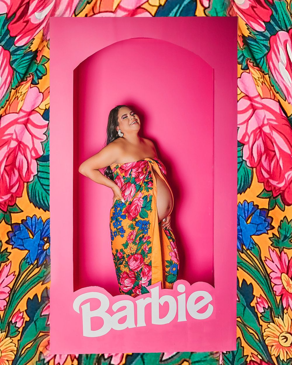 Creative maternity photo in Barbie-style box with bright pink background and floral frame.
