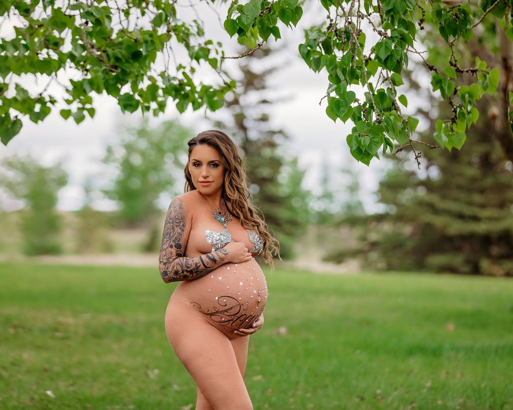 Artistic maternity photo taken outdoors under green tree branches in a natural park setting.