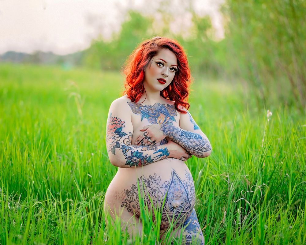 A person with vibrant red hair and tattoos poses outdoors in a field of tall green grass.