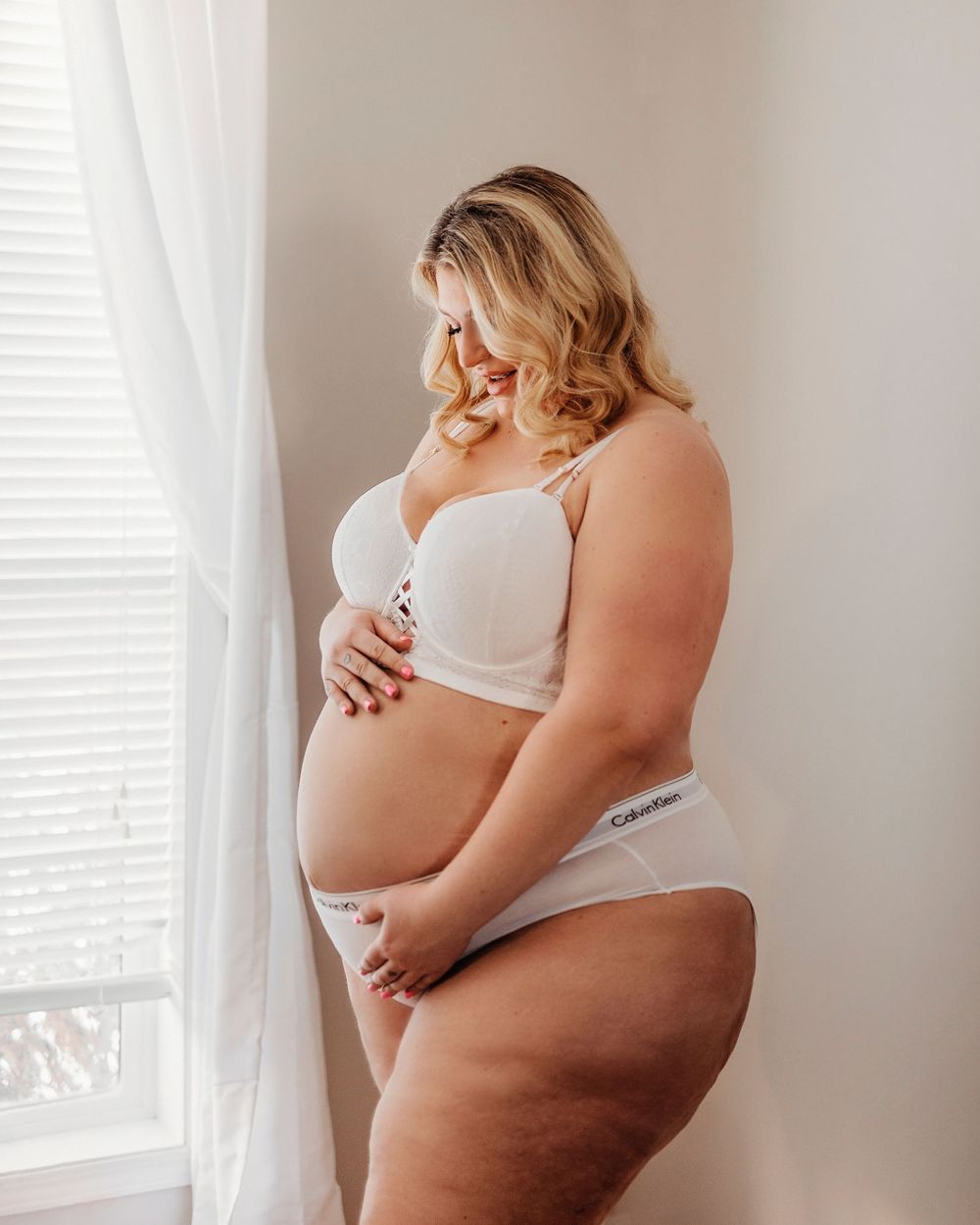 A person in white lingerie stands by a window in a soft, natural light maternity photo.