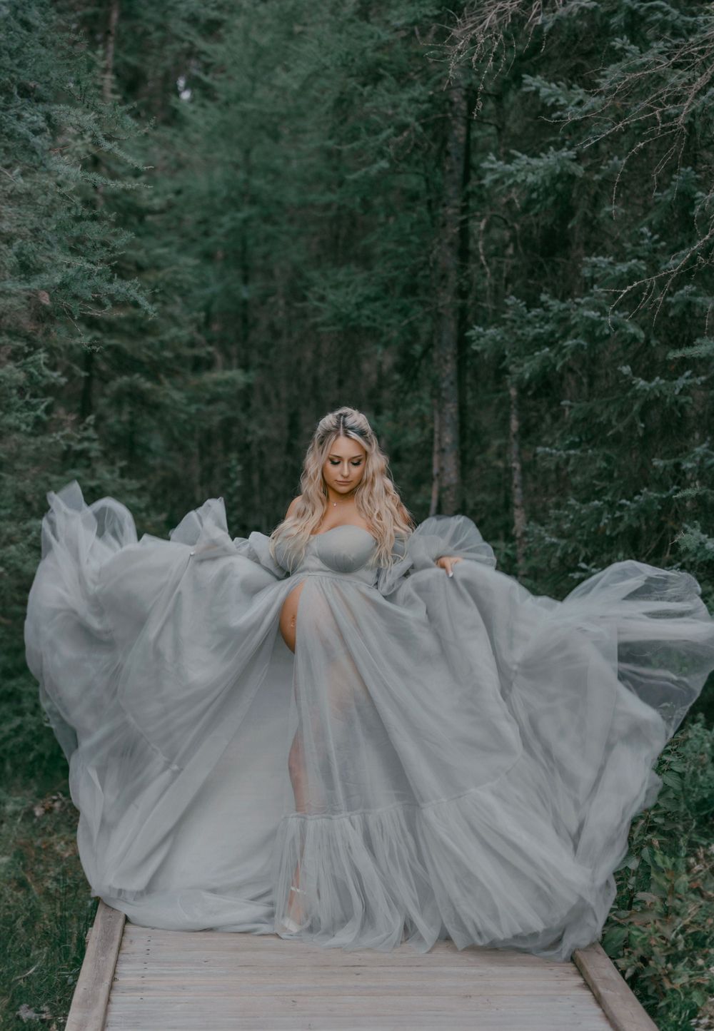 Flowing gray dress billows dramatically on wooden walkway in forest.