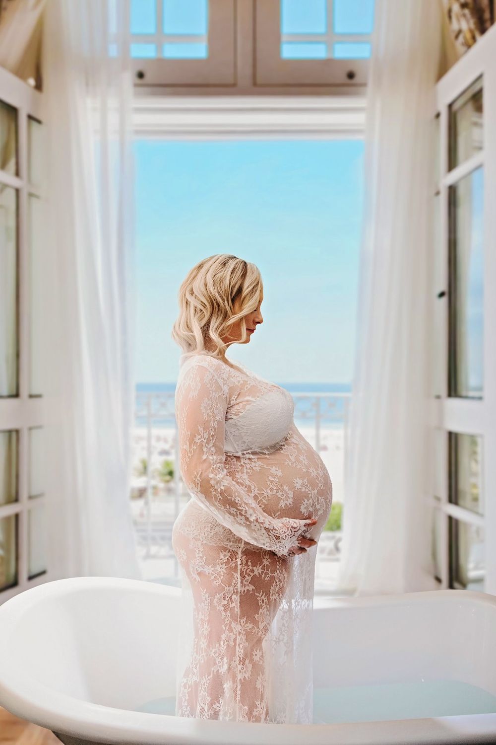 Elegant maternity photo in white lace dress by a bright window overlooking a city view.