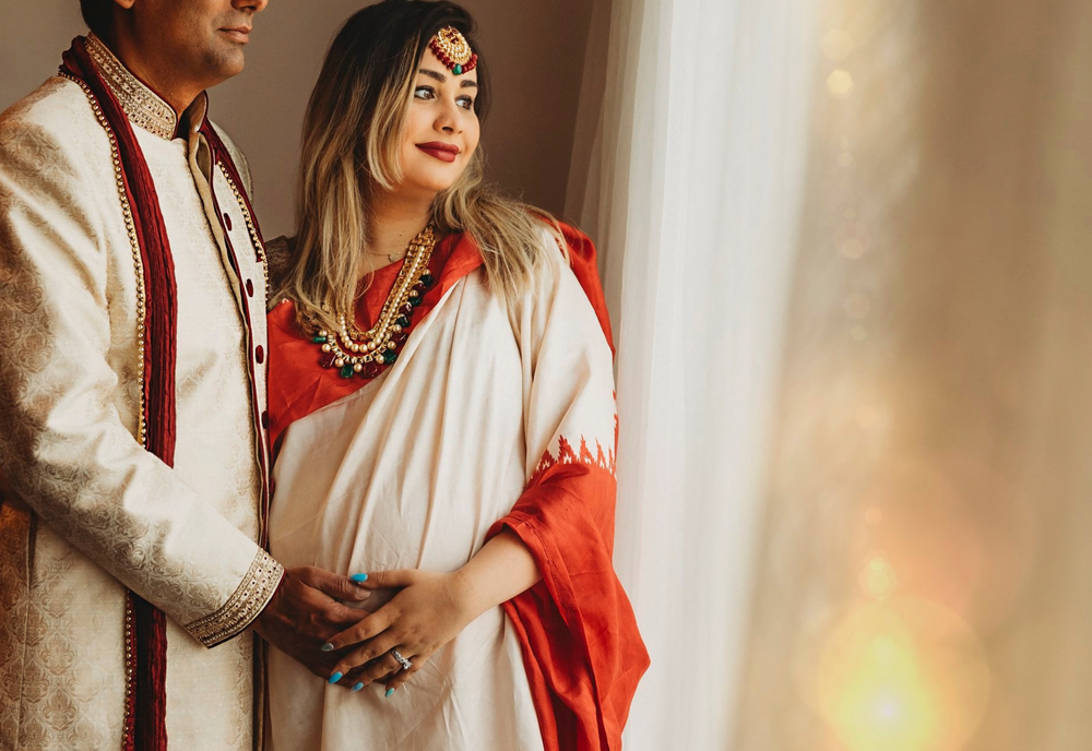 Traditional South Asian couple in wedding attire share tender moment in sunlit room.