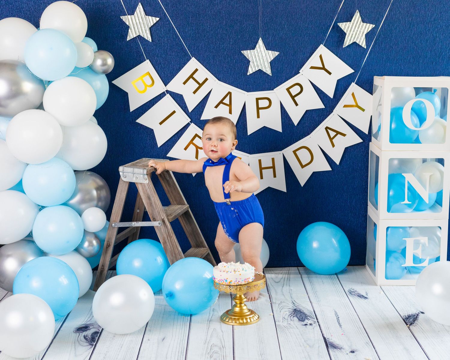 First birthday setup with blue and white balloons, ladder, and Happy Birthday banner.