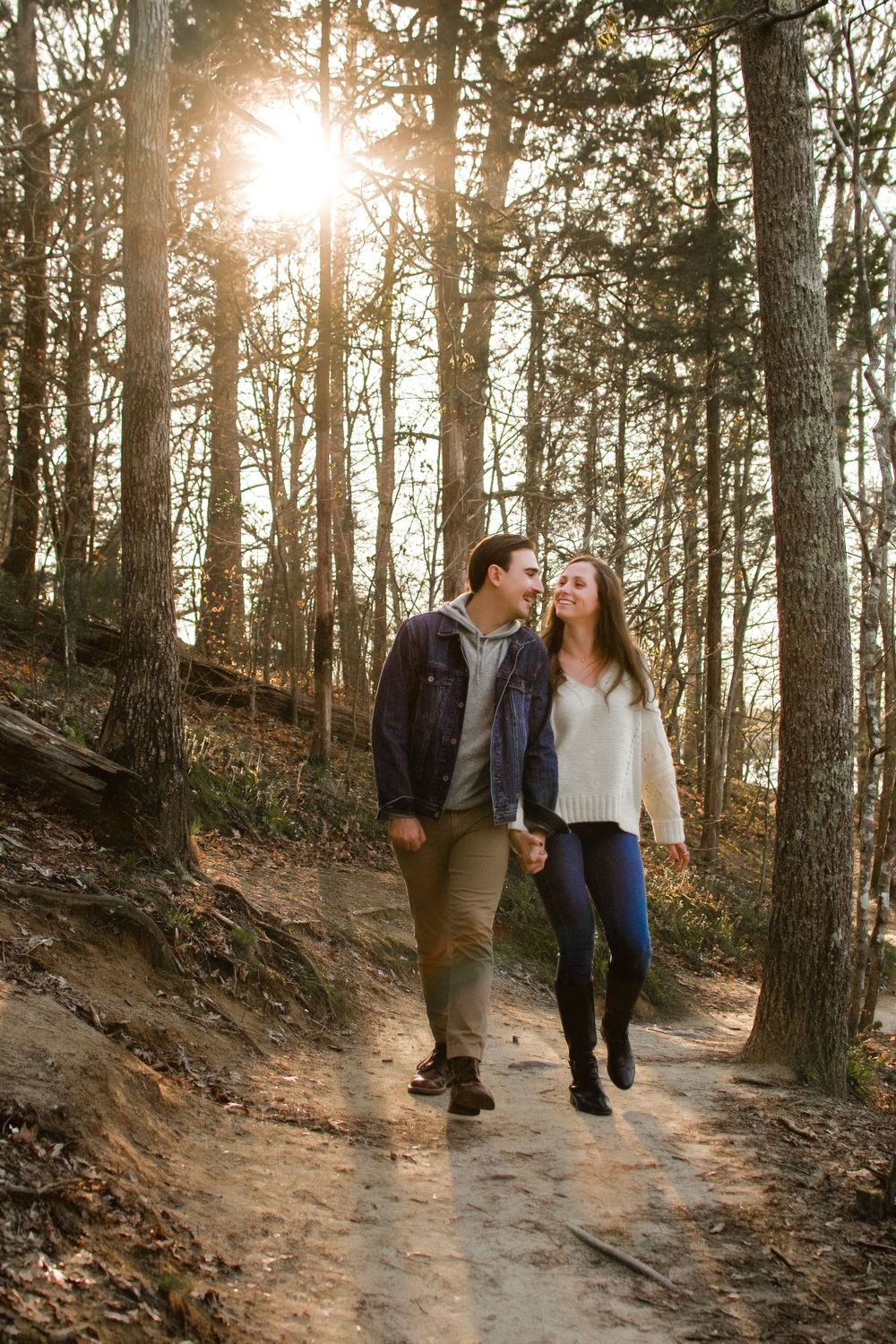 A man and a woman walking through the woods with the sunset behind them. They are laughing and looking at each other.