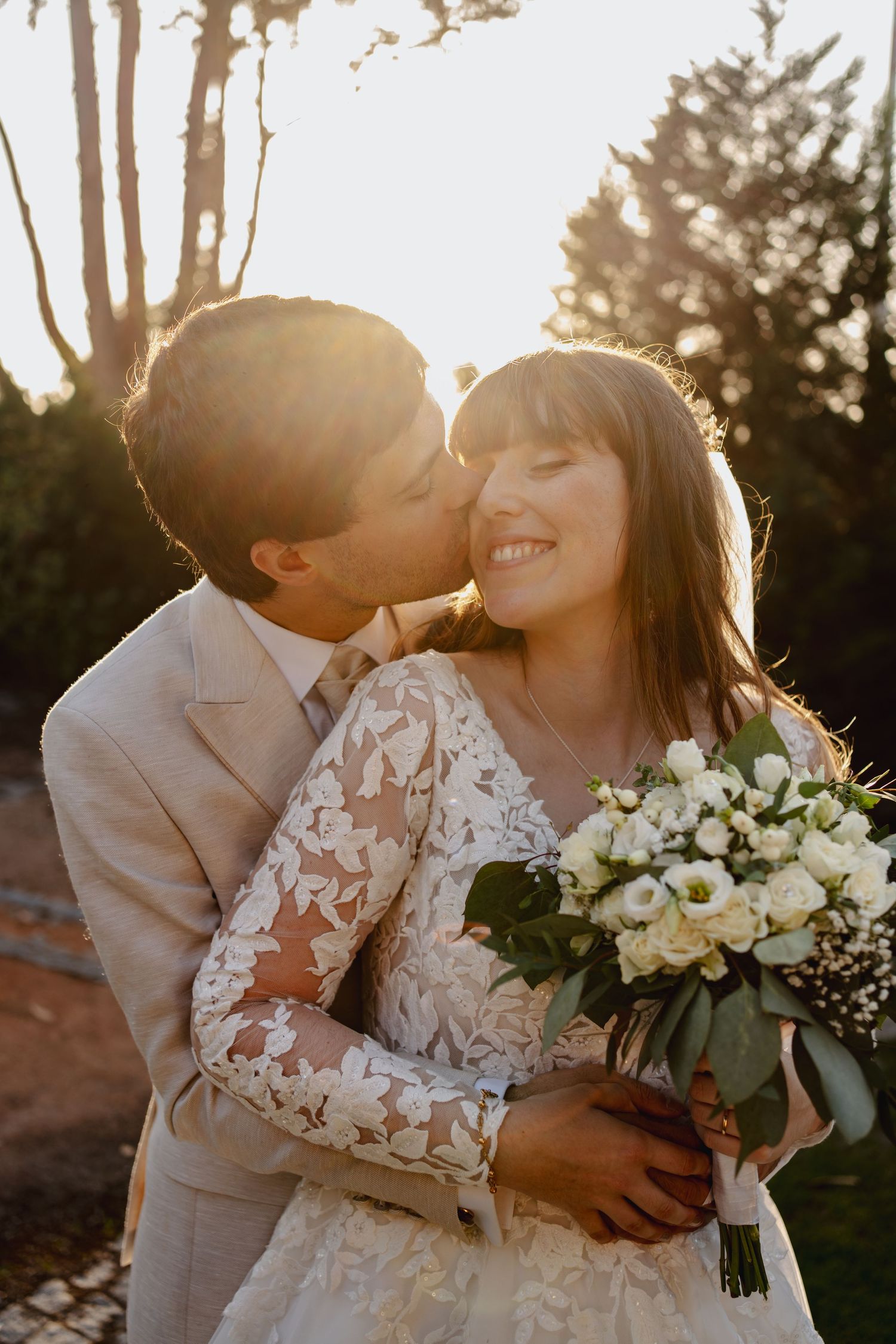 Newlyweds share intimate moment with bride holding white bouquet in golden sunset light.