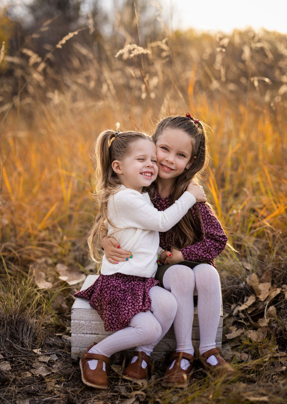 Two young children in burgundy dresses and white tights share an affectionate moment in a golden autumn field.