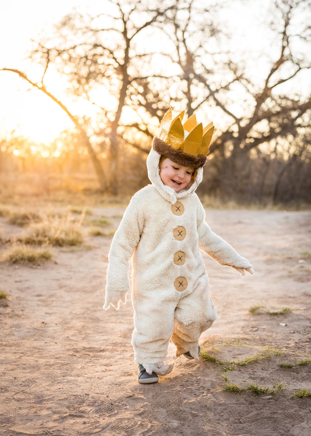 Child in white plush costume with gold crown plays on dirt path at sunset.